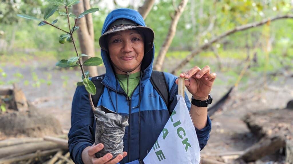 Eine sehr sympatische Frau zeigt lächelnd einen Mangroven-Setzling in die Kamera und hält eine abgeknickte Fahne von Click A Tree in ihrer anderen Hand.